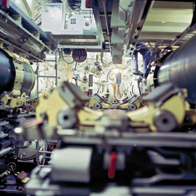 Ein Mann steht auf einer Maschine in einem industriellen Raum, umgeben von anderen Maschinen, arbeitet an der Ausrüstung.