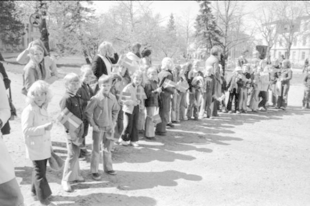 Eine Gruppe von Menschen steht in einer Reihe auf einem Schotterweg, hält Fahnen, mit Bäumen, Gebäuden und einem klaren Himmel im Hintergrund, auf einem Schwarz-Weiß-Bild einer Protestkundgebung auf dem Schulgelände.