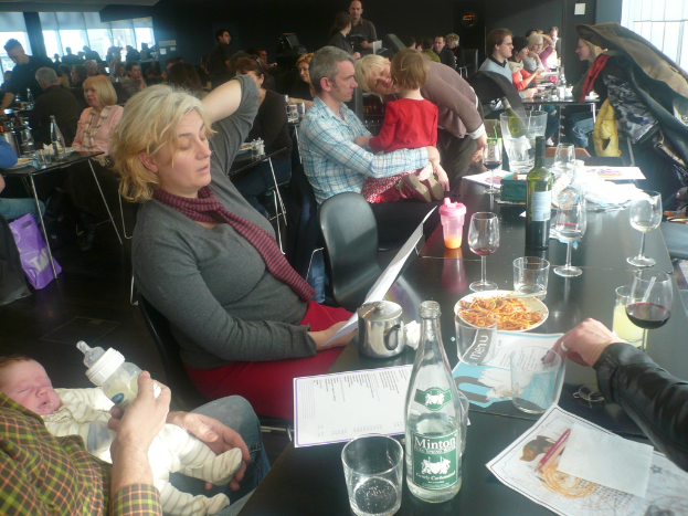 Ein Innenblick in ein belebtes Restaurant mit Gästen an Tischen, die mit Gläsern, Tellern mit Essen, Getränkeflaschen, Menükarten und anderen Gegenständen gedeckt sind.