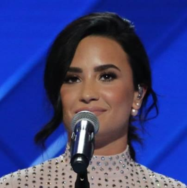 Demi Lovato steht mit einem Mikrofon auf der Bühne bei den Grammys, trägt ein schwarzes Kleid mit einem eleganten Bob, vor einem blauen Hintergrund, sieht selbstbewusst und entschlossen aus.