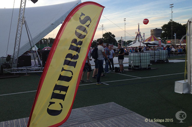 Eine belebte Karnevals Szenerie mit Zelten, einem zentralen Mast, Menschen, Bäumen, Strommasten, Straßenbahnen auf der Straße und einem Eisenständer auf der linken Seite unter einem klaren Himmel.