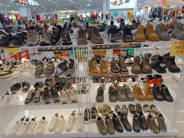 Ein überfüllter Schuhladen mit verschiedenen Schuhen auf Gestellen, Preisetiketten und Menschen im Hintergrund, mit Kleiderauslagen, Deckenlampen und einem Bildschirm sowie einem Wasserzeichen, das einen Standort in China anzeigt.