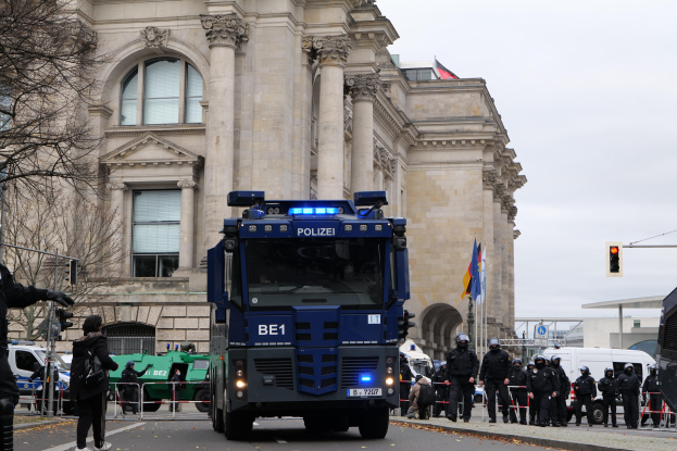Gruppe von Polizisten vor einem großen Gebäude mit Säulen und Rundbogenfenstern, mit Fahrzeugen auf der Straße und einer Person mit einer Kamera links.