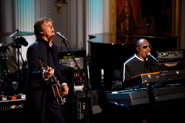 Paul McCartney spielt Gitarre und singt in ein Mikrofon, während Stevie Wonder am Piano sitzt und eine Brille trägt, gemeinsam auf den Grammys mit Instrumenten und einem gerahmten Bild im Hintergrund.