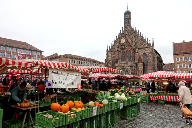 Ein belebter Markt in Nürnberg, Deutschland, mit farbenfrohen Obst- und Gemüseauslagen, Menschen mit Taschen und Zelten, vor Gebäuden mit Fenstern und einem Uhrenturm unter einem sichtbaren Himmel.