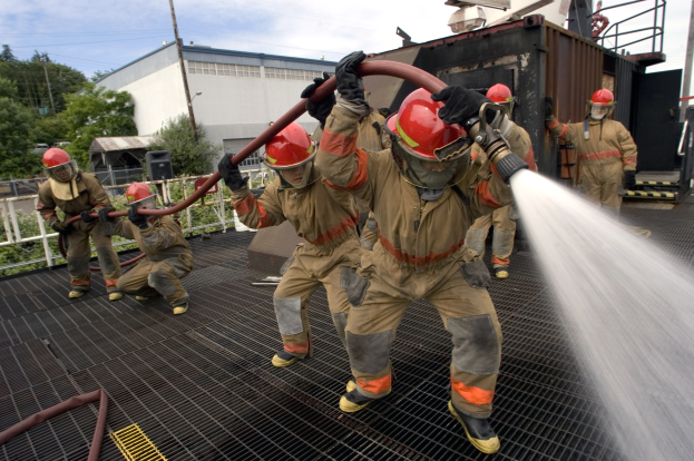 Feuerwehrleute in Helmen und Handschuhen spritzen Wasser auf ein Feuerwehrauto mit Bäumen, Gebäuden und Strommasten im Hintergrund unter einem bewölkten Himmel.