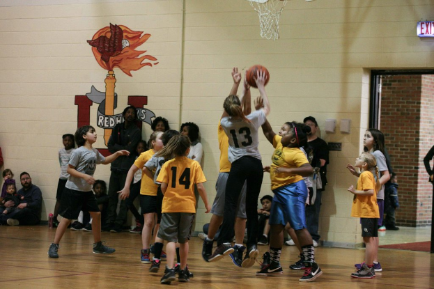 Kinder in gelben und grauen T-Shirts beim Basketballspielen, mit einem Mädchen in einem grauen T-Shirt mit der Nummer 13, das einen Wurf versucht. Im Hintergrund sitzt ein Mann in einem blauen Hemd neben einer weißen Wand mit Text, und rechts davon ist ein Ausgangsschild und ein rotes Backsteingebäude zu sehen.