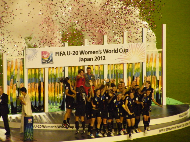 Eine Gruppe von Fussballspielerinnen in schwarzen Uniformen steht auf einer Bühne und feiert ihren Sieg, umgeben von einer Menge mit Kameras, mit einem dekorierten Hintergrund und einem grasbedeckten Boden.