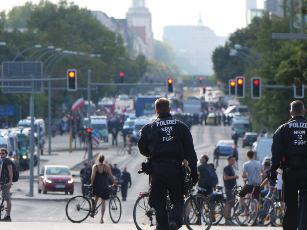 Zwei Polizeibeamte neben einer Gruppe von Radfahrern auf einer Straße mit Fahrzeugen, Verkehrszeichen, Bäumen, Gebäuden und einem klaren blauen Himmel im Hintergrund.