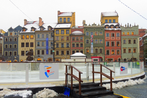 Eislaufbahn in einem Stadtplatz umgeben von Geb├Ąuden, Treppen, Gel├Ąndern, Informationsschildern, schneebedeckter Erde, Laternenmasten, B├Ąumen und einem Himmel im Hintergrund.