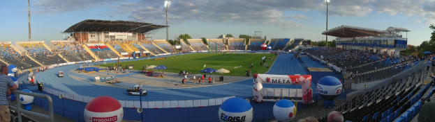 Großes Stadion mit Zuschauern bei einer Leichtathletikveranstaltung, umgeben von Bäumen, Laternenmasten, Gebäuden und Fahrzeugen unter einem bewölkten Himmel.