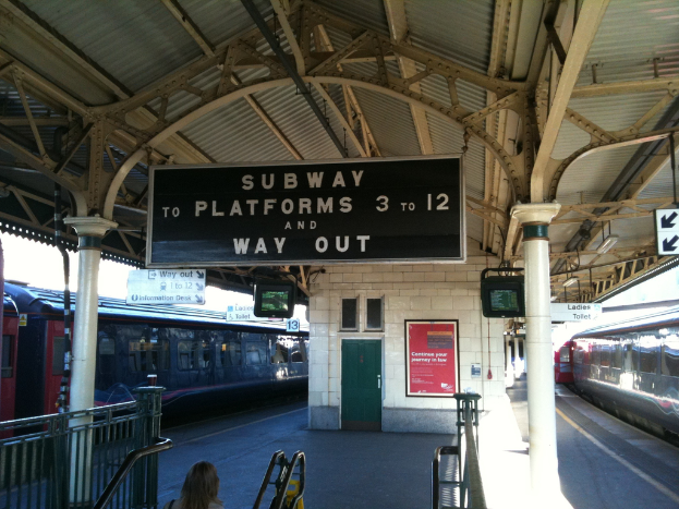 Zwei Züge stehen neben einem Bahnsteig mit Bildschirmen und Schildern, die von der Decke hängen, eine Person steht neben der Treppe und eine Tür ist an der Wand.
