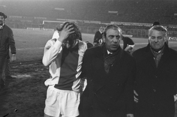 Eine Gruppe von Männern in Not steht zusammen auf einem Fußballfeld mit einem Stadion im Hintergrund, in Schwarz-Weiß aufgenommen.