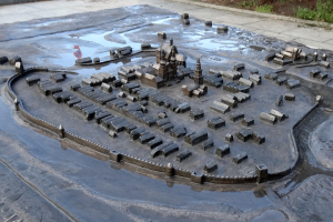 Ein Modell einer Stadt in der Mitte eines Wasserflecks, umgeben von Gebäuden, Bäumen, Pflanzen und einer Wand im Hintergrund.