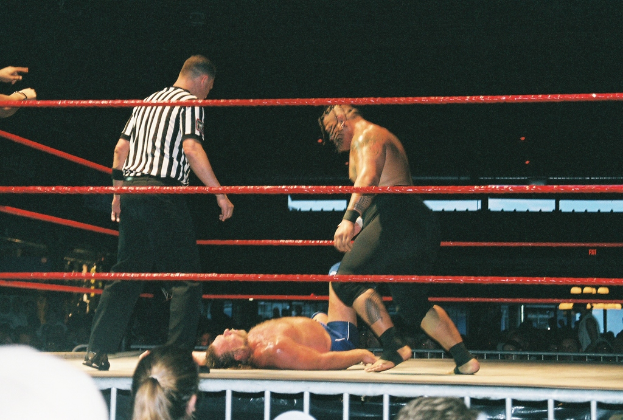 Zwei Boxer kämpfen in einem Ring mit roten Seilen, während ein Mann in einem weißen und schwarzen gestreiftem T-Shirt von der Seite zusieht.