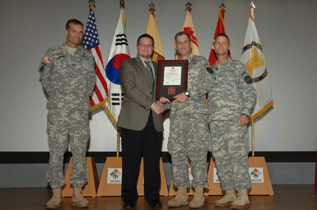 Vier Männer auf einer Bühne: drei in Armeeuniformen und einer in Blazer, Krawatte und Hemd, die Hände schütteln und einen Preis überreichen, mit sechs Flaggen auf Ständen und einer weißen Wand im Hintergrund.