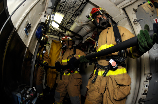 Eine Gruppe von Feuerwehrleuten in Helmen und Handschuhen hält einen Schlauch an der Rückseite eines Flugzeugs, mit Drähten, Lampen und anderen Gegenständen im Hintergrund und Text am unteren Rand des Bildes.
