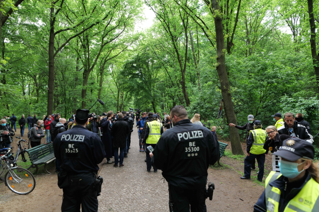 Eine Gruppe von Polizeibeamten vor einer Menge während einer Anti-Terror-Demonstration in Berlin, mit Fahrrädern und einer Bank im Vordergrund und Bäumen im Hintergrund.