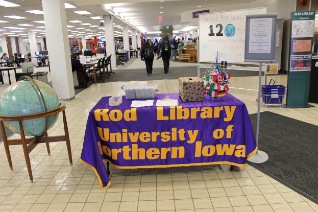 Eine Ausstellung mit einem Tisch, auf dem verschiedene Landesflaggen, eine Kiste, Papiere und ein Banner liegen, eine Kugel zur Linken, ein Anschlagbrett zur Rechten mit einigen Menschen daneben und mehreren Tischen und Stühlen links.