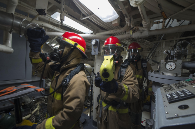 Drei Feuerwehrleute in Helmen und Gasmasken stehen vor einer Steuerkonsole, rechts daneben eine Maschine und im Hintergrund Rohre, Lampen und andere Gegenstände.