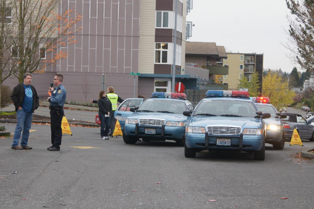 Autos auf einer Straße mit vier Menschen in der Nähe, Gebäuden mit Fenstern im Hintergrund, Bäumen und Warnkegeln.