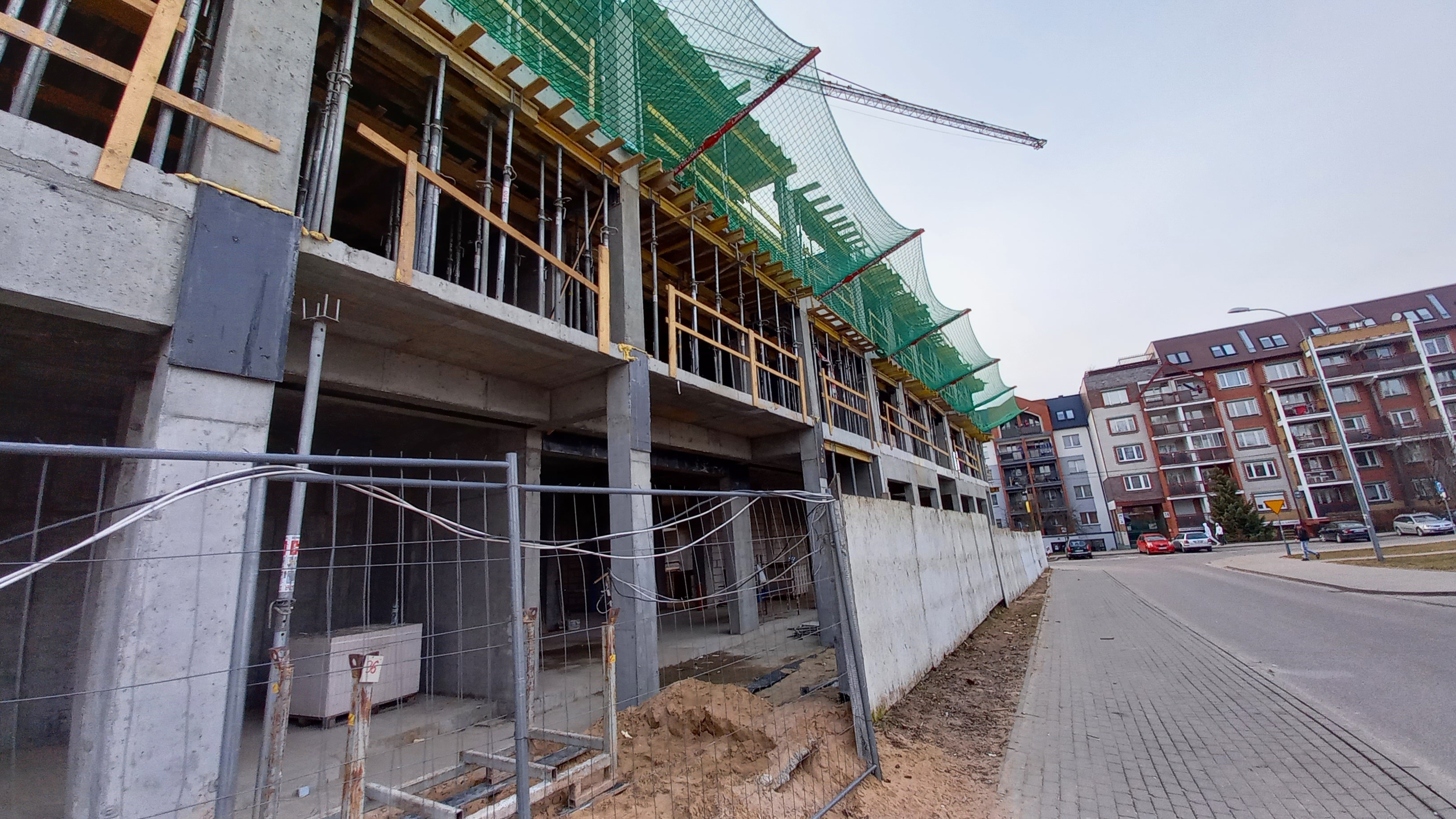 Baustelle mit einem großen Gebäude im Bau neben einer Straße, umgeben von Gebäuden, Straßenmasten, Straßenlaternen und Kraftfahrzeugen, unter einem klaren blauen Himmel.