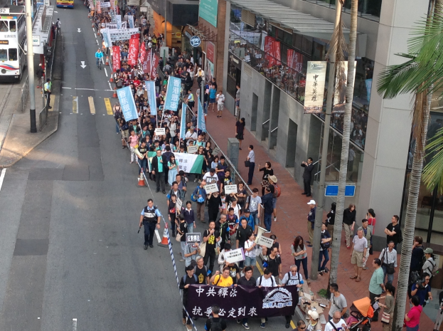 Eine große Gruppe von Menschen marschiert auf einer Straße in Hong Kong, hält Schilder und Plakate, mit Bäumen, Gebäuden und Fahrzeugen im Hintergrund.