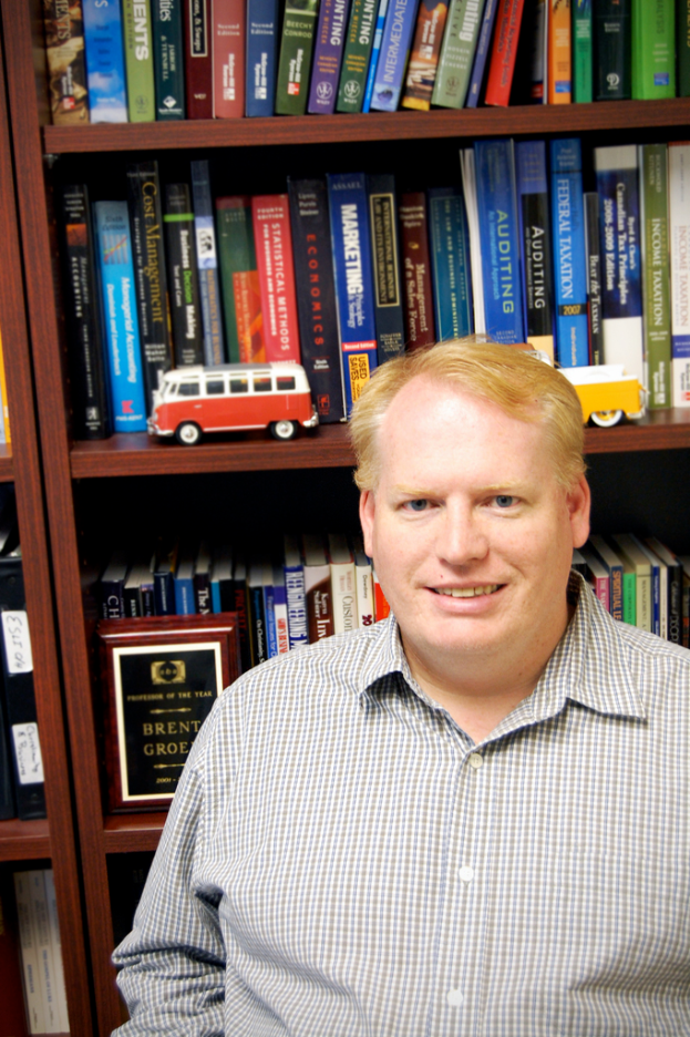 Ein Mann lacht in einem Raum mit Bücherregalen und zwei Spielzeugen im Hintergrund.