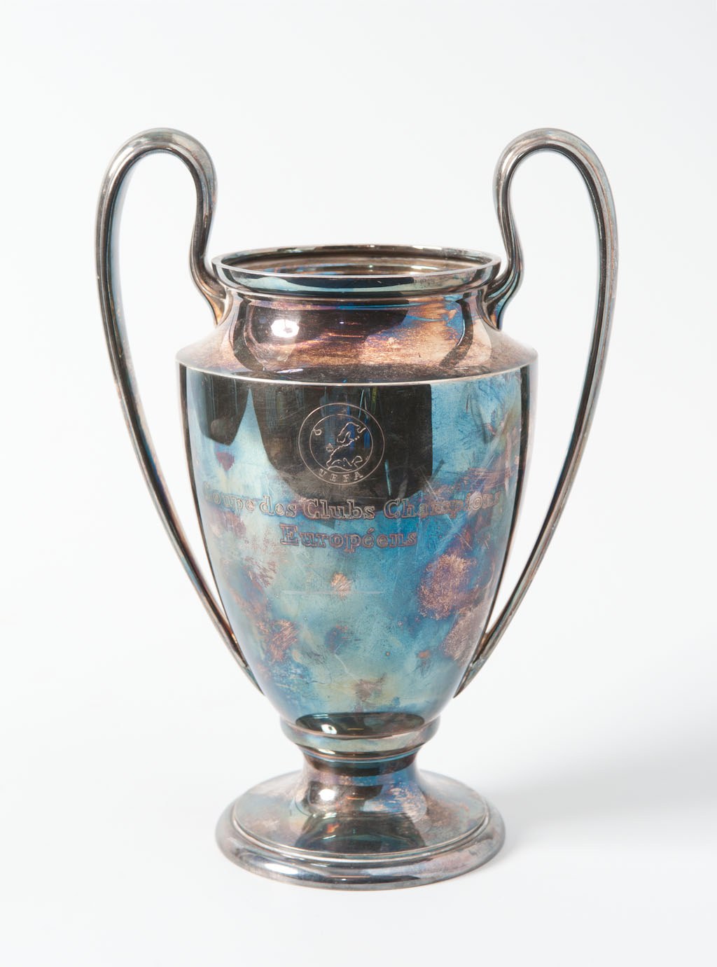 Der UEFA-Champions-League-Pokal, ein stahlerner Becher mit zwei Griffen und einem blauen und silbernen Farbschema, ist mit 'UEFA Champions League' eingraviert und wird gegen einen weißen Hintergrund dargestellt.