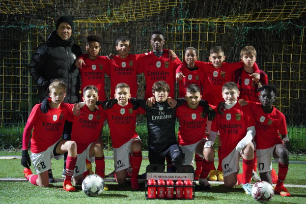 Eine Gruppe junger Jungs in roten T-Shirts und schwarzen Shorts posiert für ein Foto auf einem Fußballfeld, umgeben von mehreren Fußballbällen, einem Netz, einem Zaun und Bäumen im Hintergrund.