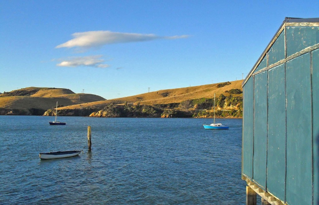 Boote und Pfähle im Wasser, ein Holzhaus auf der rechten Seite, Bäume und Berge mit Windmühlen im Hintergrund und Wolken am Himmel.