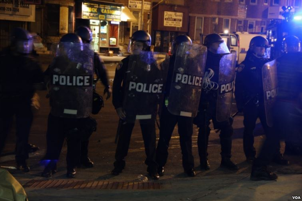 Eine Gruppe von Polizisten in Helmen und Schilden steht in einer Straße bei Nacht, mit Gebäuden, Fahrzeugen und Laternen im Hintergrund.
