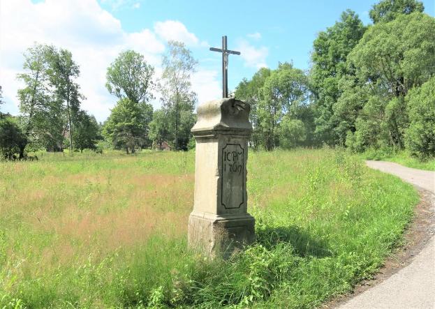 Ein Steinkreuz in einer gr"{u}nen Wiese neben einer Stra{\ss}e, umgeben von B"{a}umen und einem bew"{o}lktem Himmel, dient als Holocaust-Gedenkst"{a}tte mit eingraviertem Text.