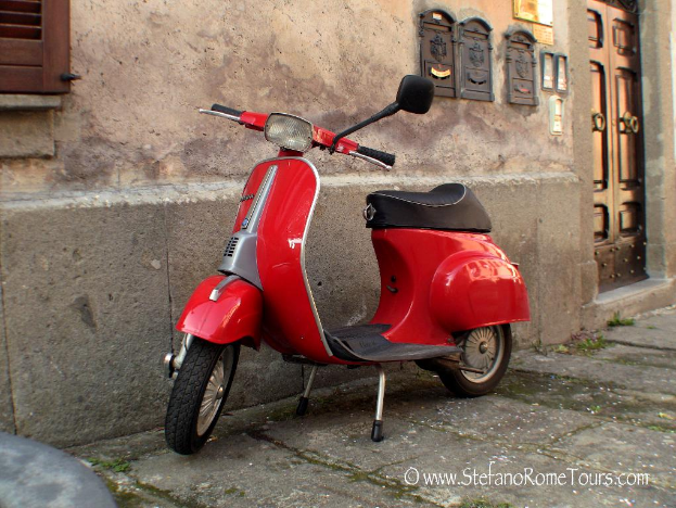 Ein roter E-Scooter steht vor einem Haus mit Tür und Fenstern auf einer Straße mit lesbarer Schrift.