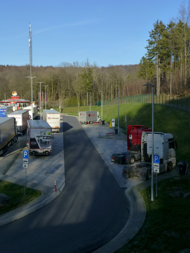 Mautstelle mit mehreren Lastwagen, die an der Straße geparkt sind, Laternenmäste, Schilder, Verkehrskegel, Gras, ein Zaun, Bäume und ein Turm im Hintergrund bei klarem blauem Himmel.