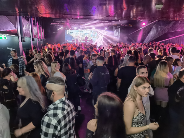 Eine große Gruppe von Menschen in einer Diskothek mit Deckenbeleuchtung und Säulen, mit einem Bildschirm im Hintergrund, der ein Absolut Nightlife-Event anzeigt.