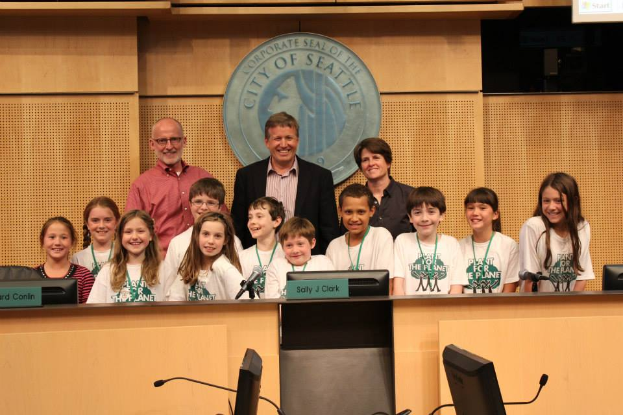 Eine Gruppe von Kindern und Erwachsenen steht lachend vor einem Rednerpult, mit Monitoren, Mikrofonen und anderen Gegenständen auf einem Tisch im Vordergrund, einem Logo an der Wand und einem Bildschirm auf der rechten Seite, was auf eine Stadtratssitzung hindeutet.