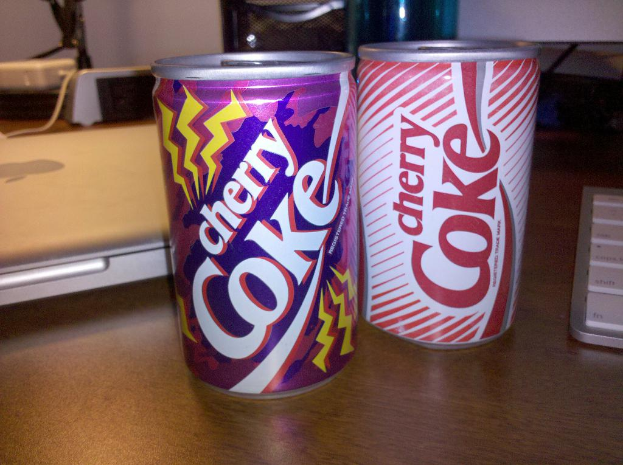 Zwei Coke-Dosen, eine Tastatur und ein Laptop sind auf einem Tisch platziert.