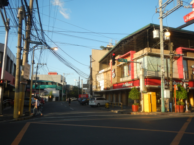 Eine Stadtstraße mit Autos, Fußgängern, Strommasten, Laternen, Ampeln, Gebäuden mit Schildern, Topfpflanzen und einer bewölkten Himmel.