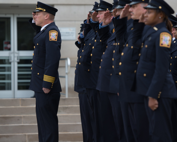 Eine Gruppe von Polizisten in Uniform und Mützen, die vor einem Gebäude mit Glastüren und Treppen salutieren.