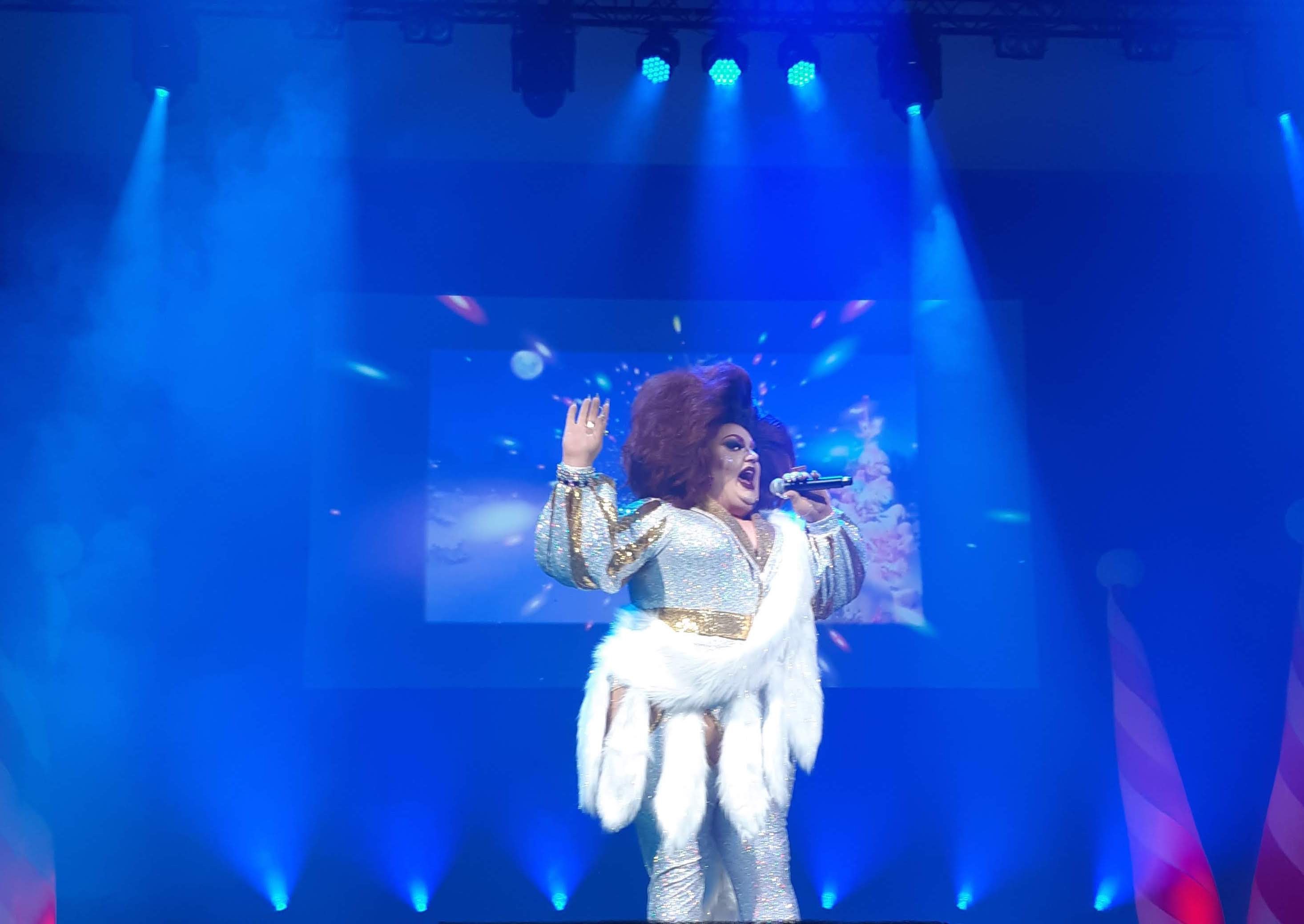 Frau in weißem Kleid singend auf einer Bühne mit hellen Scheinwerfern und einem Bildschirmhintergrund, hält ein Mikrofon während RuPaul's Drag Race All Stars.