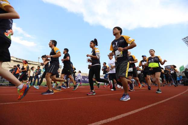 Gruppe von Läufern auf einer Laufbahn mit Zuschauern auf der rechten Seite, Gebäuden und Bannern im Hintergrund unter einem klaren blauen Himmel.