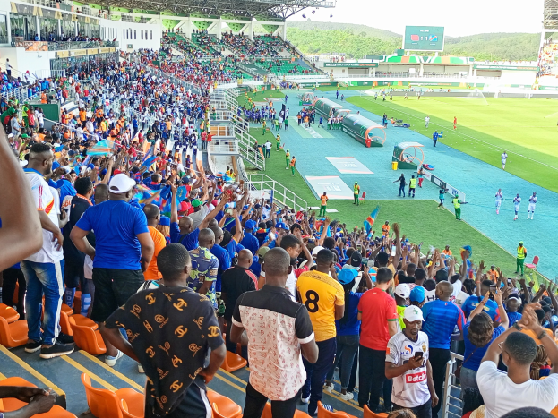 Eine große Menschenmenge versammelt sich vor einem eingezäunten Fußballfeld, mit einigen auf Stühlen sitzend und anderen stehend, mit Blick auf Hügel unter einem klaren blauen Himmel während der 2019 African Cup of Nations.