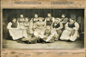 Eine Gruppe von Frauen in traditioneller russischer Kleidung posiert für ein Foto, einige sitzen auf dem Boden und andere stehen, vor einem Vorhanghintergrund, mit Text oben und unten auf dem Bild.