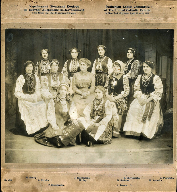Eine Gruppe von Frauen in traditioneller russischer Kleidung posiert für ein Foto, einige sitzen auf dem Boden und andere stehen, vor einem Vorhanghintergrund, mit Text oben und unten auf dem Bild.