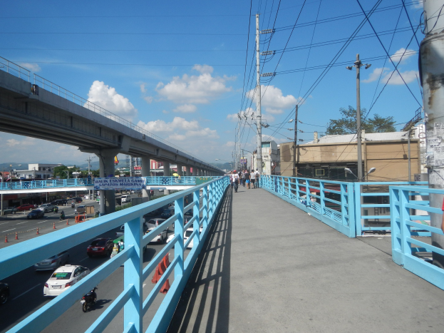 Eine Fußgängerbrücke mit Menschen darauf, darunter Fahrzeuge und im Hintergrund Gebäude, Bäume und Himmel, mit einer Person auf der rechten Seite.