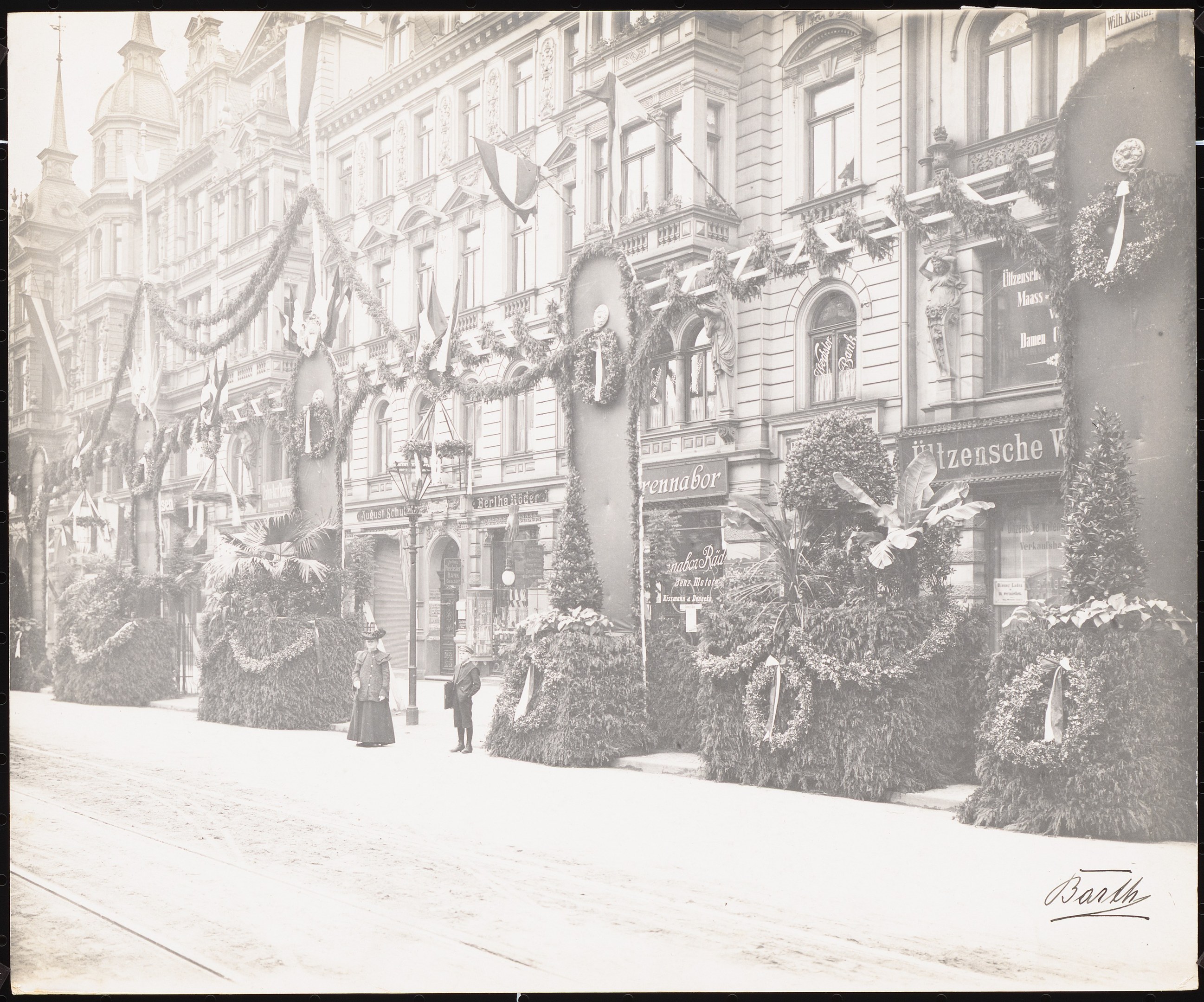 Altes Schwarz-Weiß-Foto einer Stadtstraße mit Fußgängern, Gebäuden mit Fenstern, Pflanzen, Fahnen und Namensschildern sowie einer Straße unten.