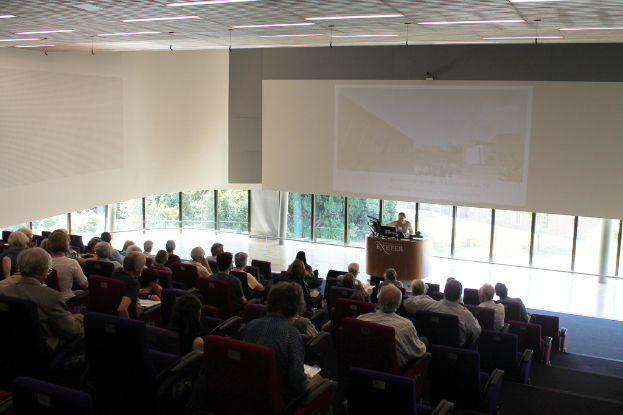 Ein Hörsaal an einer Universität mit vielen Sitzenden, die einem Präsentator an einem Tisch mit einer Präsentation gegenübersitzen, und großen Fenstern, die Bäume und Gebäude im Hintergrund zeigen.