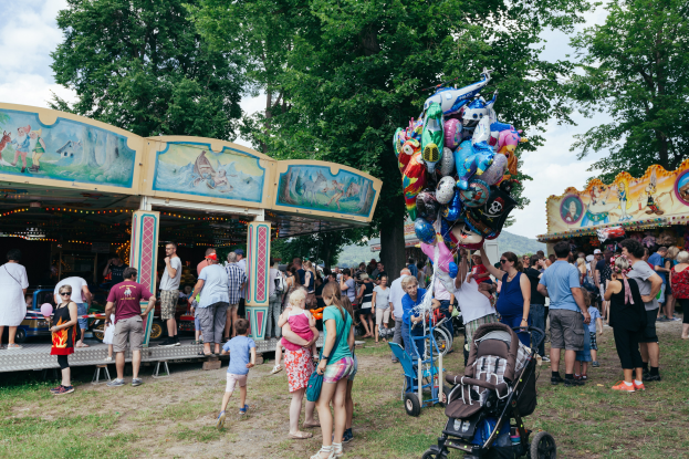 Eine Menschenmenge umgibt einen bunt beleuchteten Karussellwagen auf einem Volksfest, einige halten Kinderwagen, mit Bäumen und einem bewölktem Himmel im Hintergrund.