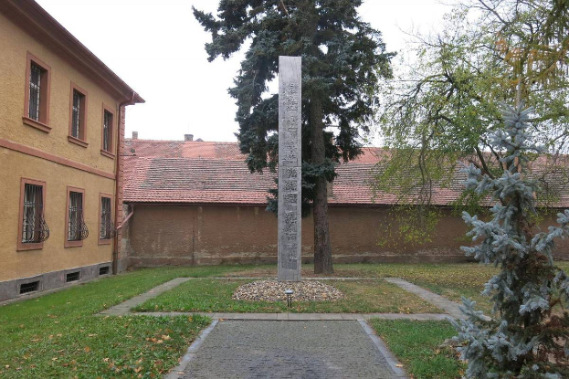Ein Denkmal steht auf einer Rasenfläche neben einem Baum, umgeben von Gebäuden mit Fenstern, einem Pfad und trockenen Blättern auf dem Boden, unter einem bewölkten Himmel und ruft eine traurige Atmosphäre hervor, die an ein Auschwitz-Mahnmal erinnert.